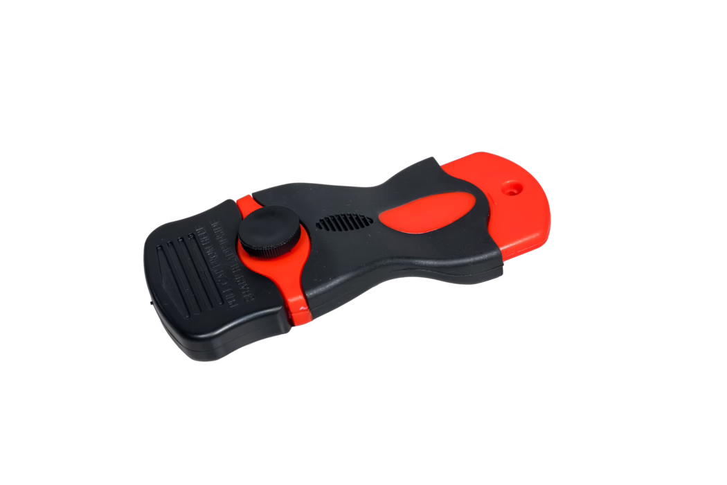 A handheld plastic utility tool in black and red photographed on a white background. The ergonomic, contoured body is mostly matte black with red accent panels and a red sliding end piece with a small hanging hole. A round textured adjustment knob, a vent-like slot, and a ridged grip area are visible; the tool appears to be a safety cutter or scraper with a concealed blade compartment. The object is shown centered and slightly rotated, with even studio lighting and a clean high-contrast backdrop.