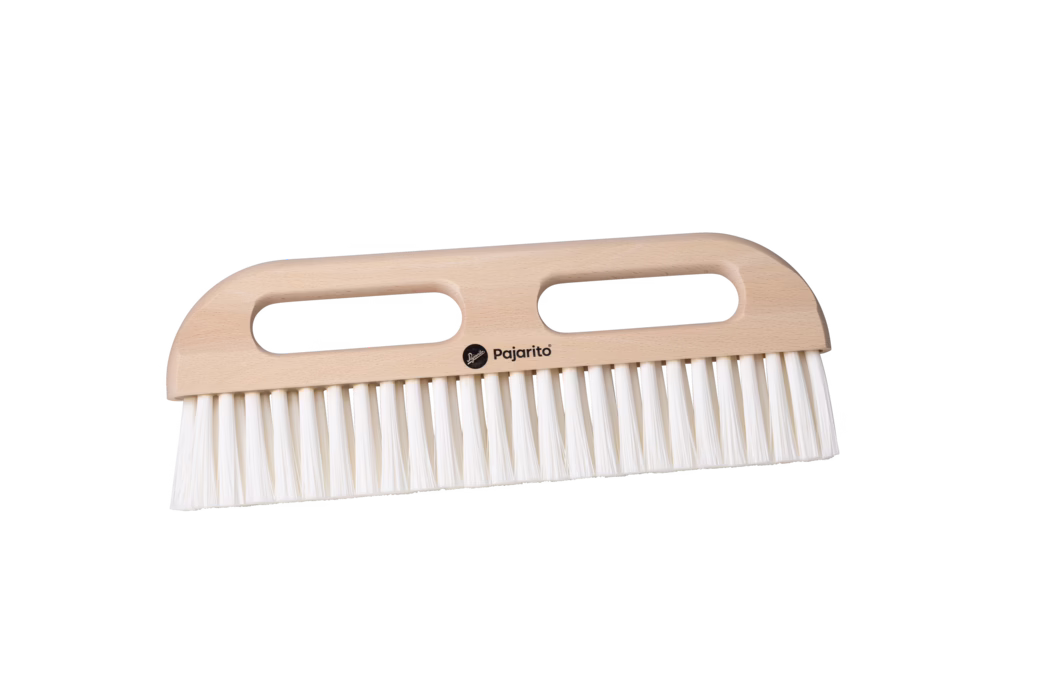 Pajarito_Dummy_Wallpaper squeegee nylon P-Line_179112_2024-02 Close-up product photo of a wooden hand brush with two elongated oval hand openings and a smooth light beech wood handle. The brush features dense, straight white synthetic bristles arranged in a single row along the lower edge. A small black 'Pajarito' logo is printed at the center of the handle. Photographed on a clean white background with soft, even lighting, shown in a horizontal orientation, suitable for household sweeping and dusting.