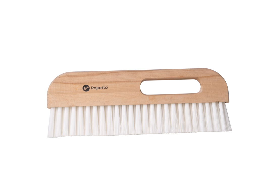 Close-up of a handheld wooden brush with a rectangular light-wood handle and an oval finger hole, showing the brand name 'Pajarito' printed on the handle; dense white synthetic bristles extend straight from the base. The brush is placed horizontally on a neutral white background and appears clean and unused. Designed for dusting and sweeping small debris from furniture and floors, it combines a smooth natural wood grip with resilient bristles for everyday cleaning.