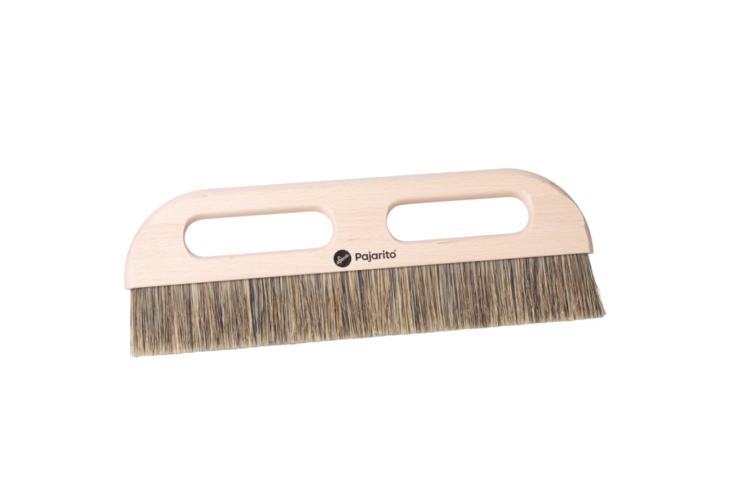 Pajarito_Dummy_Wallpaper squeegee grey bristle P-Line_179113_2024-02 Close-up of a wooden hand brush with a light beech wood handle and dense natural bristles. The flat rectangular handle features two elongated oval hand grips for a secure two-handed hold and a small black Pajarito logo printed at the center. The bristles consist of mixed tan and brown fibers, trimmed evenly to a straight edge and suitable for sweeping fine dust and small debris from floors or work surfaces. The brush is photographed on a clean white background, showing the smooth sanded wood grain, rounded corners and practical ergonomic design.